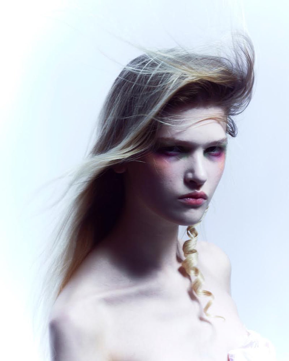 Portrait of a young woman with blonde hair and vibrant makeup, posing in a photo studio. The image features soft lighting and a clean background.