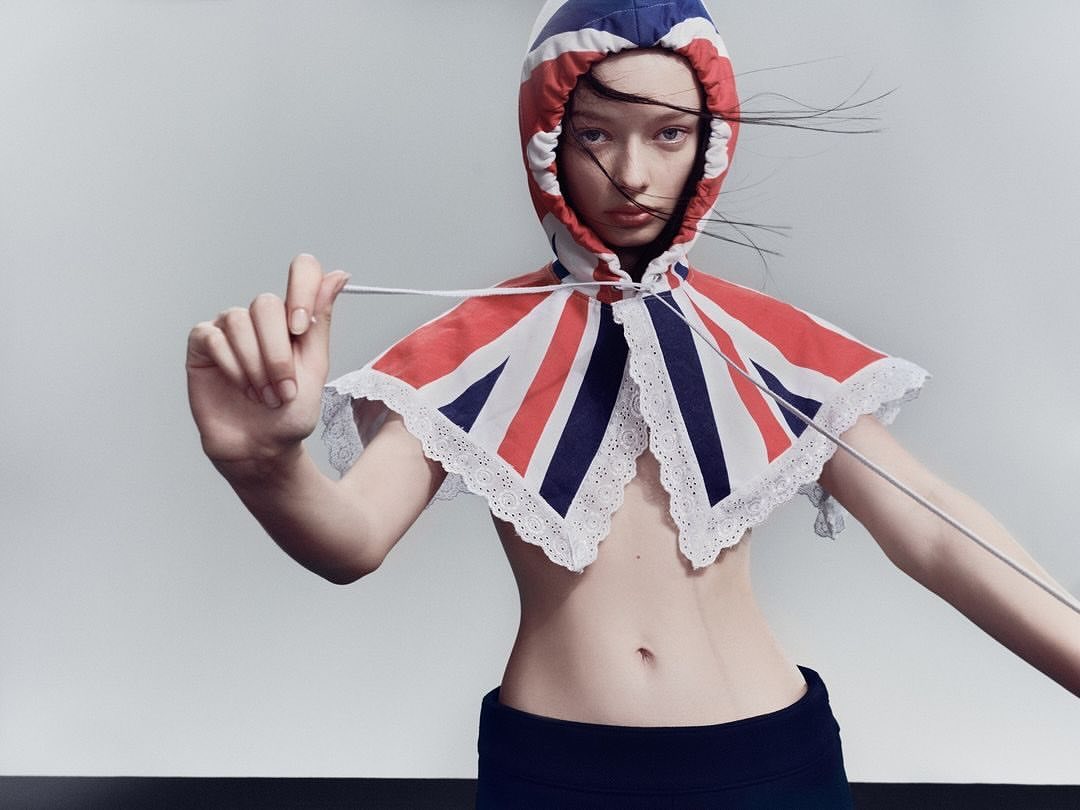 A young fair-skinned woman is seen in a studio shot, wearing a British flag-themed hooded shawl, posing in front of a grey backdrop with a black floor.