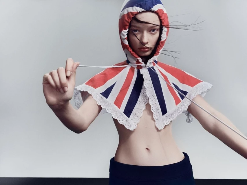 A young fair-skinned woman is seen in a studio shot, wearing a British flag-themed hooded shawl, posing in front of a grey backdrop with a black floor.