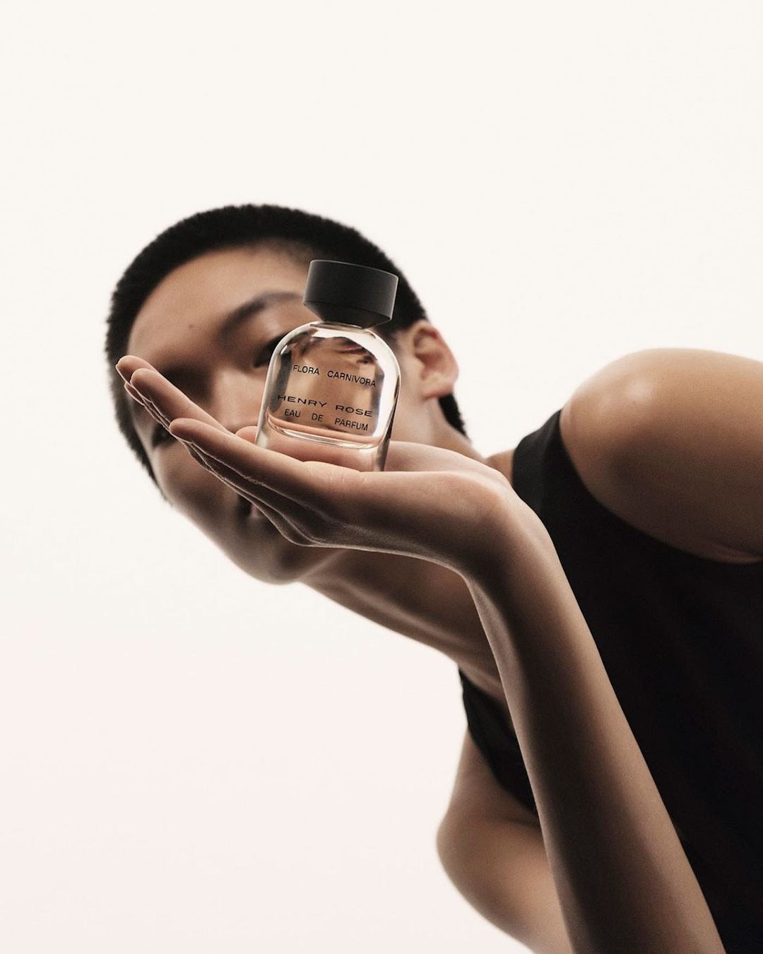 Person holds Henry Rose eau de parfum. Low angle shot captures the clear glass bottle, black cap, and neutral skin tones against a white background.