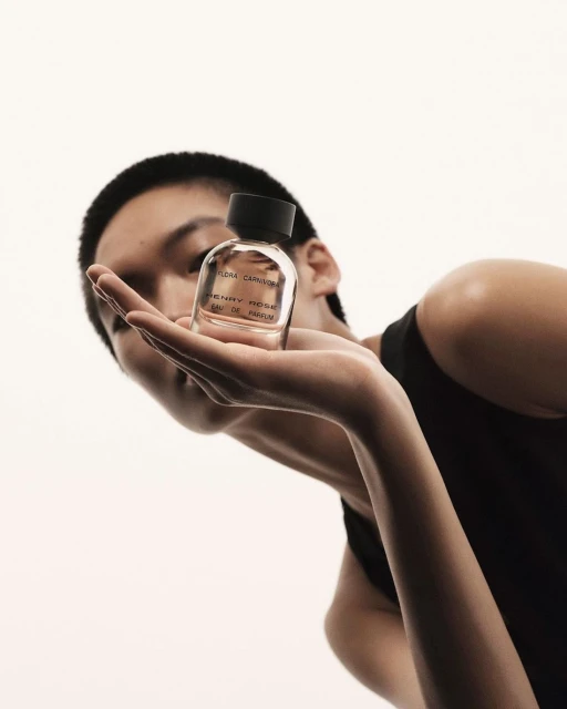 Person holds Henry Rose eau de parfum. Low angle shot captures the clear glass bottle, black cap, and neutral skin tones against a white background.