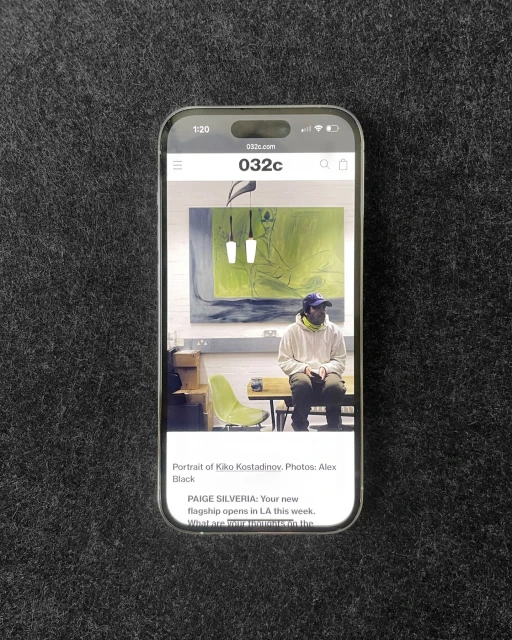 Smartphone displays an image of a person in streetwear, seated indoors. An abstract painting hangs on the wall behind them.