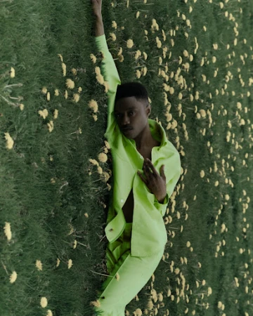 An adult male laying in a field of white and yellow flowers, dressed in a lime-green pantsuit and open dress shirt.