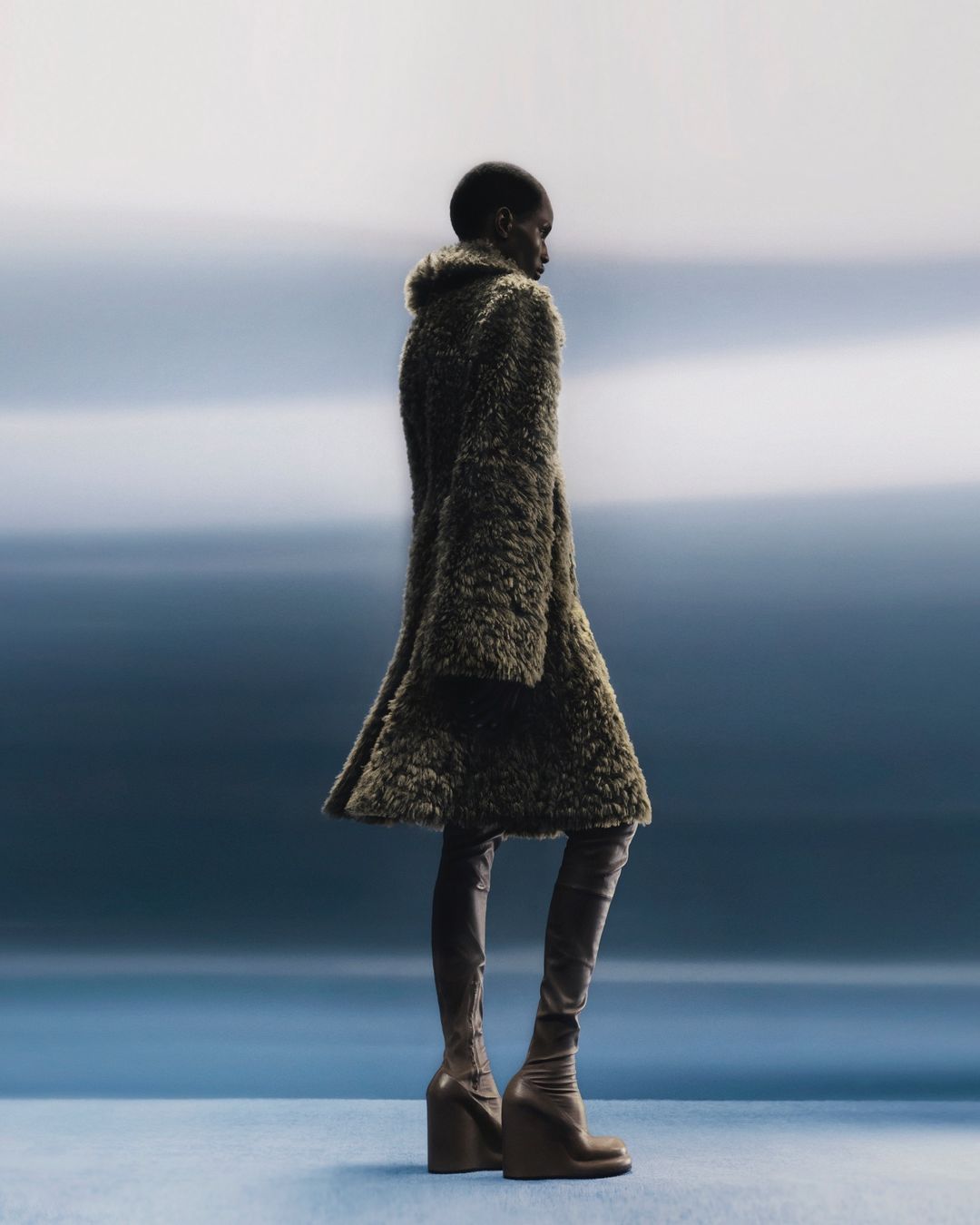 Fashion model posing in a furry olive green coat and matching brown thigh high wedge boots on a blue gradient background in a studio setting.