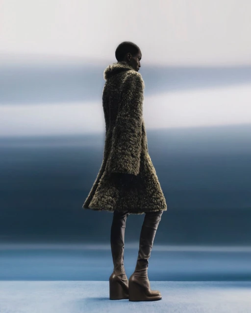 Fashion model posing in a furry olive green coat and matching brown thigh high wedge boots on a blue gradient background in a studio setting.
