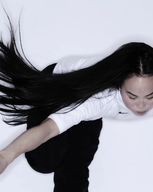 Top-down shot of a young adult woman with long black hair kneeling on a white background, wearing a white t-shirt and black pants.