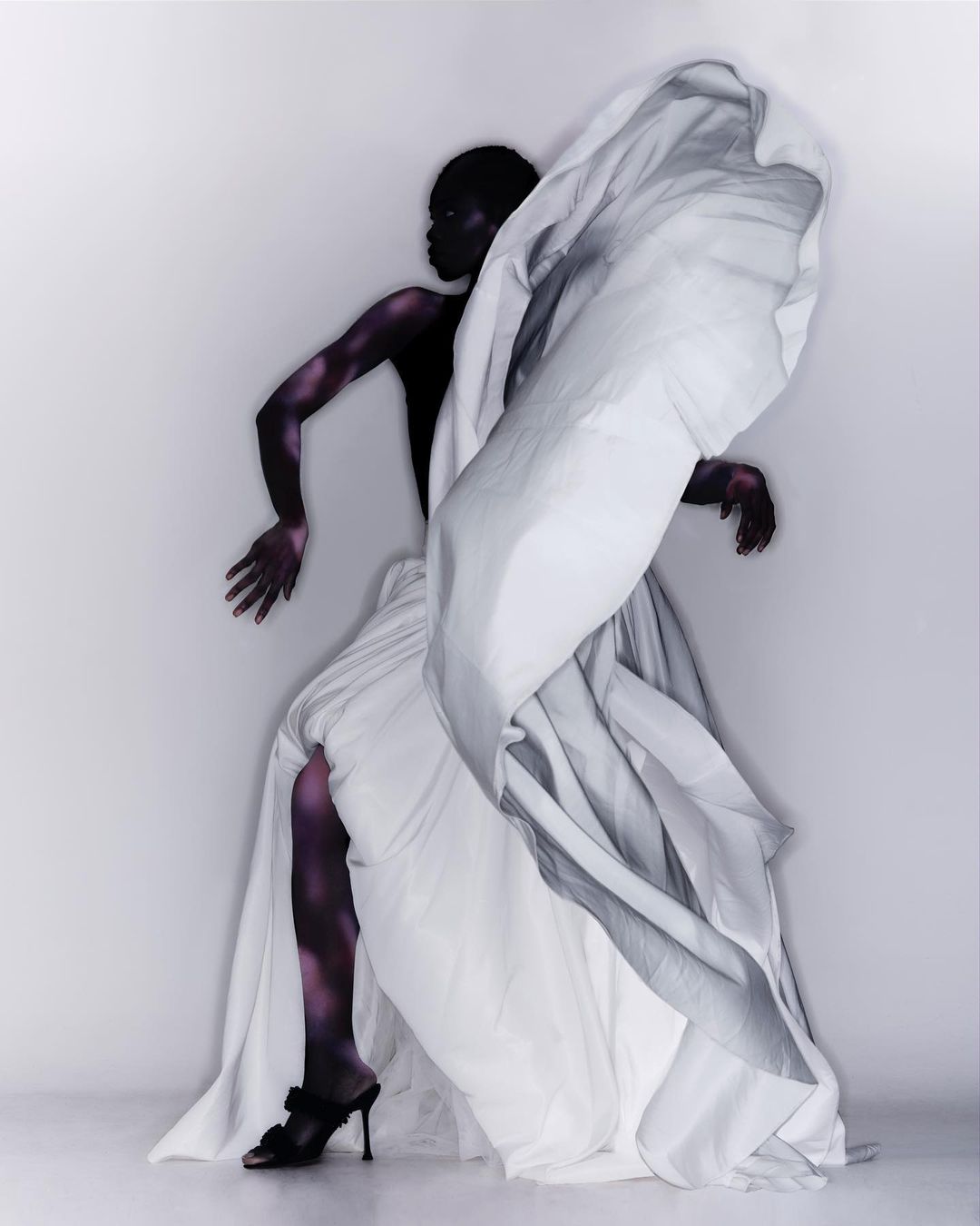 A slender woman with dark skin poses dynamically in a photo studio, draped in a flowing white dress with high-heeled mules.