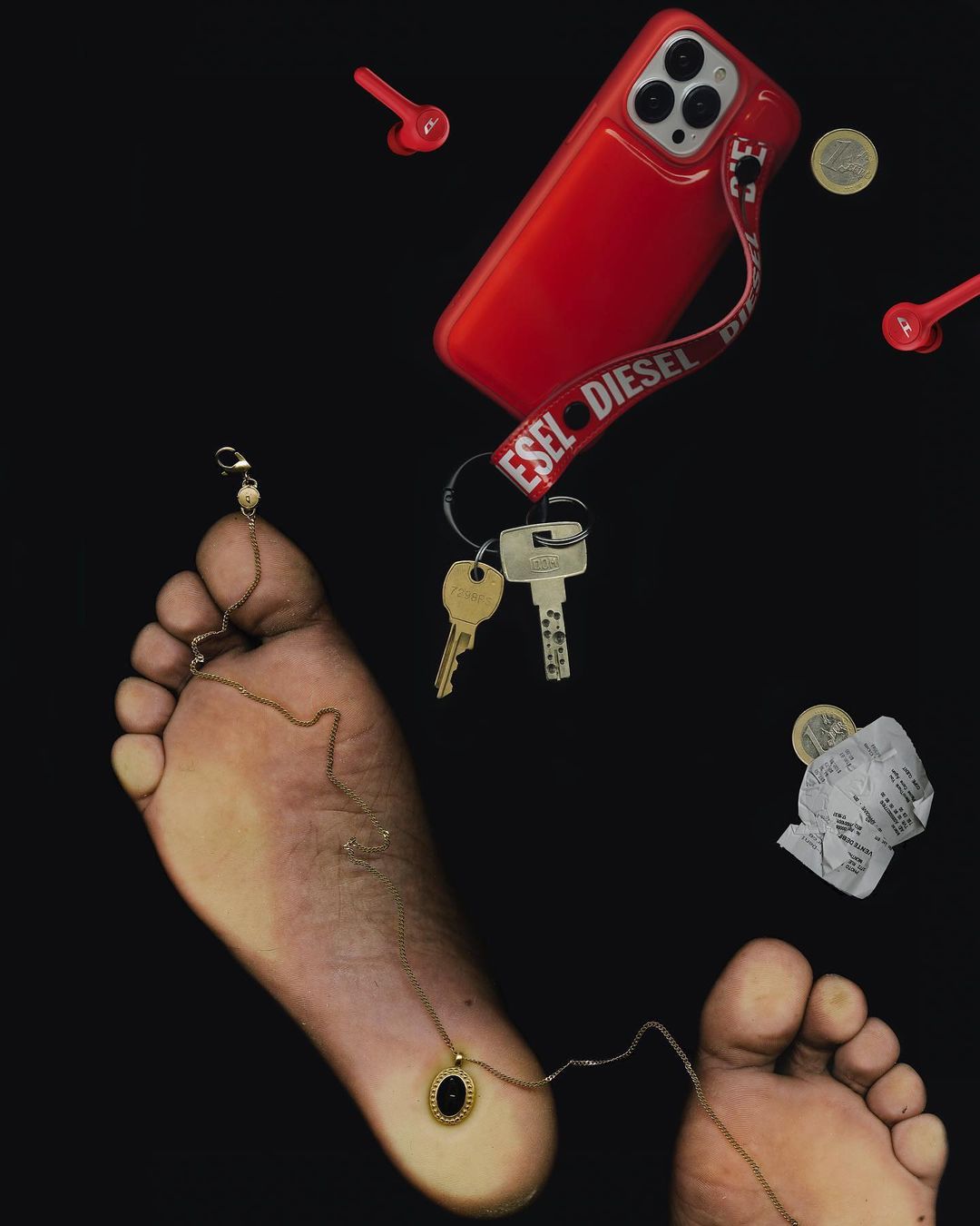 Still life composition with human feet, cell phone in red case, red earbuds, keys, jewelry and money in a dark studio setting.