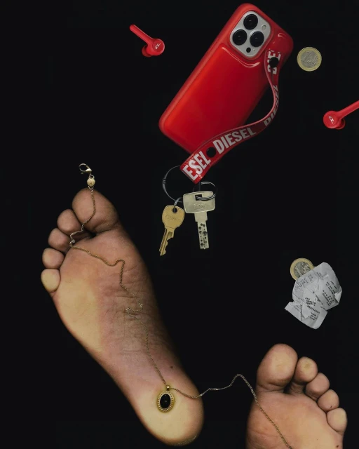 Still life composition with human feet, cell phone in red case, red earbuds, keys, jewelry and money in a dark studio setting.