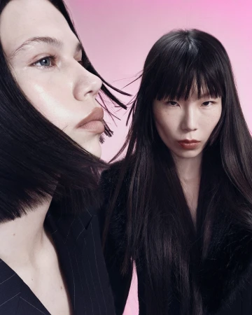 Two women with long dark hair posing against a pink gradient backdrop, wearing dark tailored blazers in a close-up beauty portrait.
