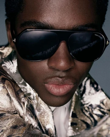 Close-up portrait of a Black man wearing oversized black sunglasses and a metallic patterned jacket.