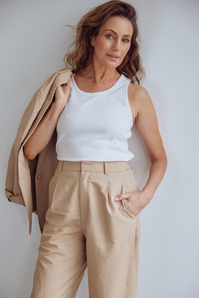 Stylish brunette woman posing in studio with white tank top, tan wide-leg pants, and a blazer draped over her shoulder, medium shot.