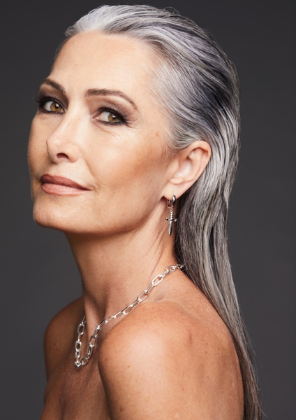 Portrait of an adult woman with gray hair styled back, wearing silver cross earrings and a matching silver necklace, against gray background.