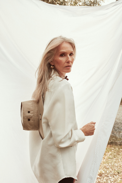 Elegant mature woman with long grey hair, dressed in an off-white jacket, poses outdoors against a draped white cloth backdrop.