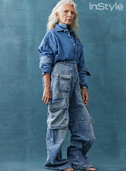 Elderly woman in denim shirt and embellished baggy jeans posing in front of a teal background for an InStyle fashion editorial shot.