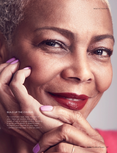 Close-up of a mature adult female showcasing red lipstick and lilac nail polish, with her hands gently framing her face in an indoor setting.