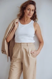 Mature woman wearing a white tank top and beige trousers holding a blazer over her shoulder against a white wall.