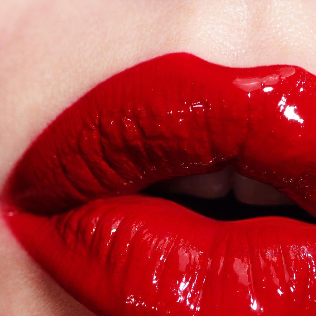 Close-up shot of a woman's face featuring glossy red lips with a full pout and visible white teeth, conveying bold beauty.