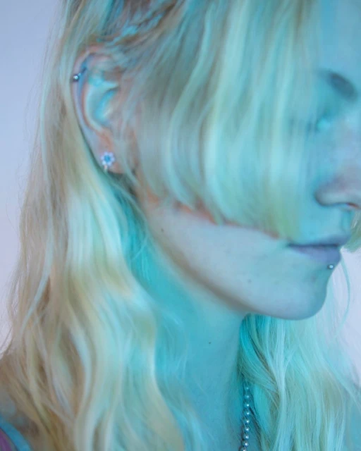 Close-up of a light-skinned female's face in a side view, showcasing blonde wavy hair, a lip piercing, and jewelry under blue lighting.