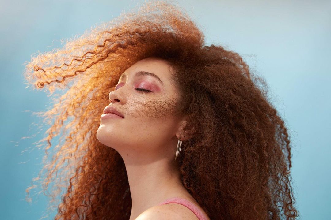 Close-up beauty shot of a woman with freckles, pink eyeshadow, and curly auburn hair, posing in front of a light blue background.