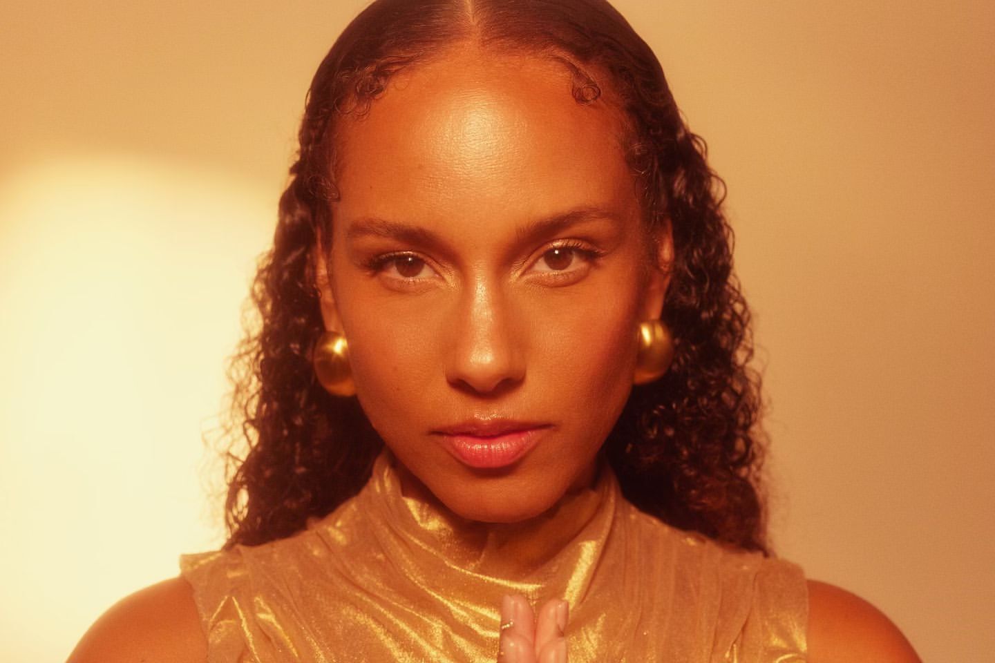 Close-up portrait of a woman with curly hair wearing gold earrings and a gold top, bathed in warm golden light in an indoor setting.