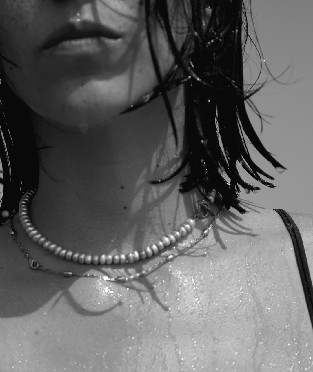 Monochrome close-up of a woman's upper body with wet hair, a pearl necklace, and water droplets on her skin in an outdoor setting.