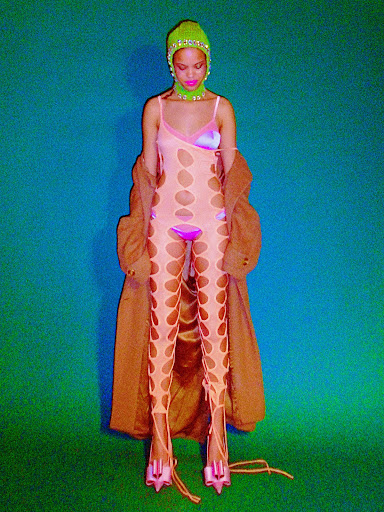 A fashion photo features a slender woman in a cut-out bodysuit, embellished hat, and high heels against a vibrant, contrasting background.
