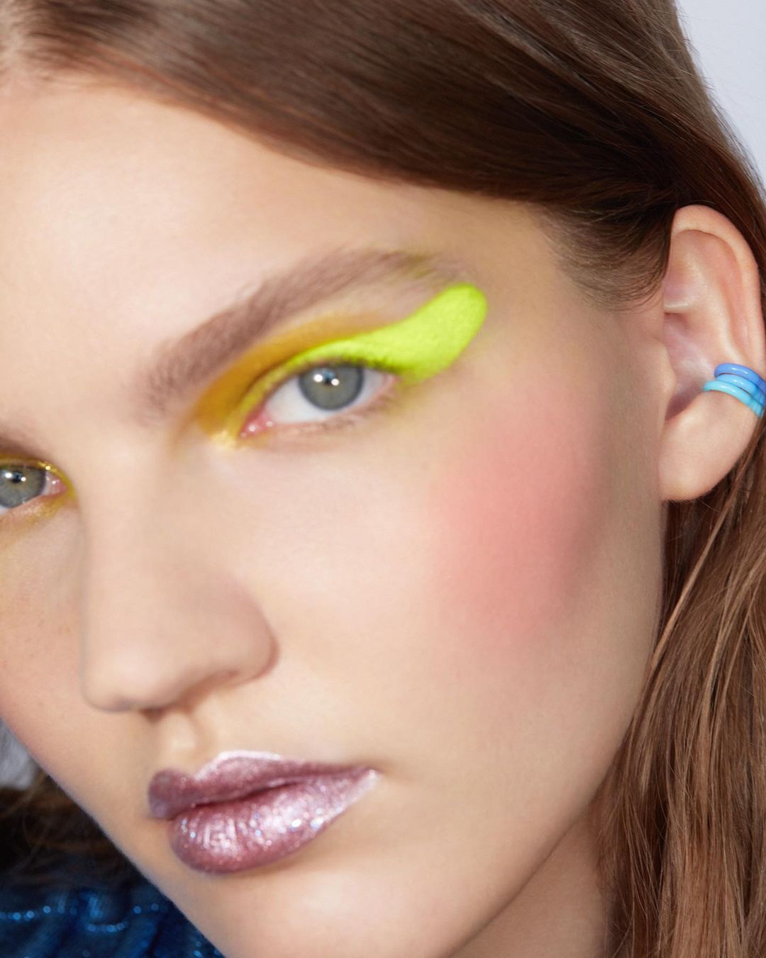 Close-up portrait of a model with vibrant neon yellow eye makeup and glossy purple metallic lipstick by Anairam.