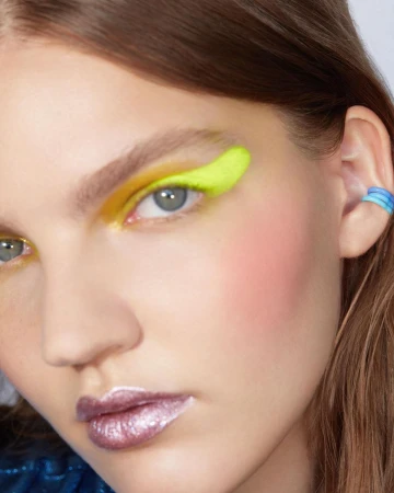 Close-up portrait of a model with vibrant neon yellow eye makeup and glossy purple metallic lipstick by Anairam.