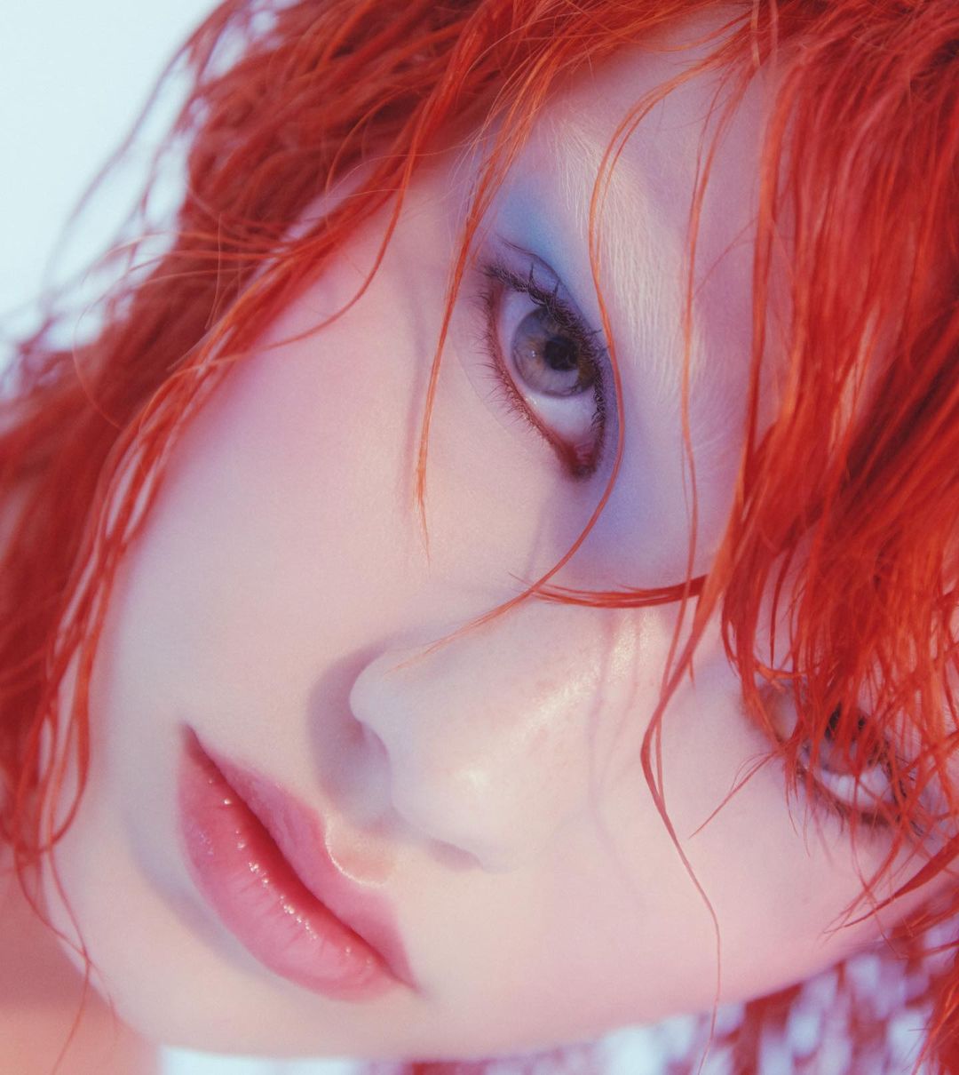 Close-up portrait of a woman with bright red hair and blue eyeshadow. Soft, diffused lighting enhances her features in a studio environment.
