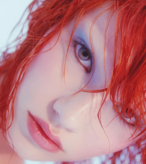 Close-up portrait of a woman with bright red hair and blue eyeshadow. Soft, diffused lighting enhances her features in a studio environment.