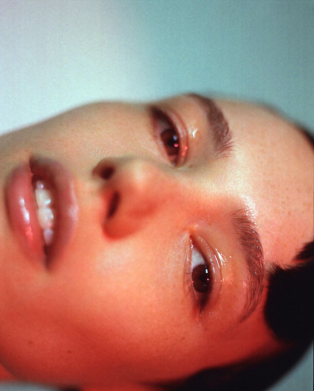 Close-up studio portrait of a young woman with brown eyes and brown hair. Her face is illuminated with red light, and skin is dewy.