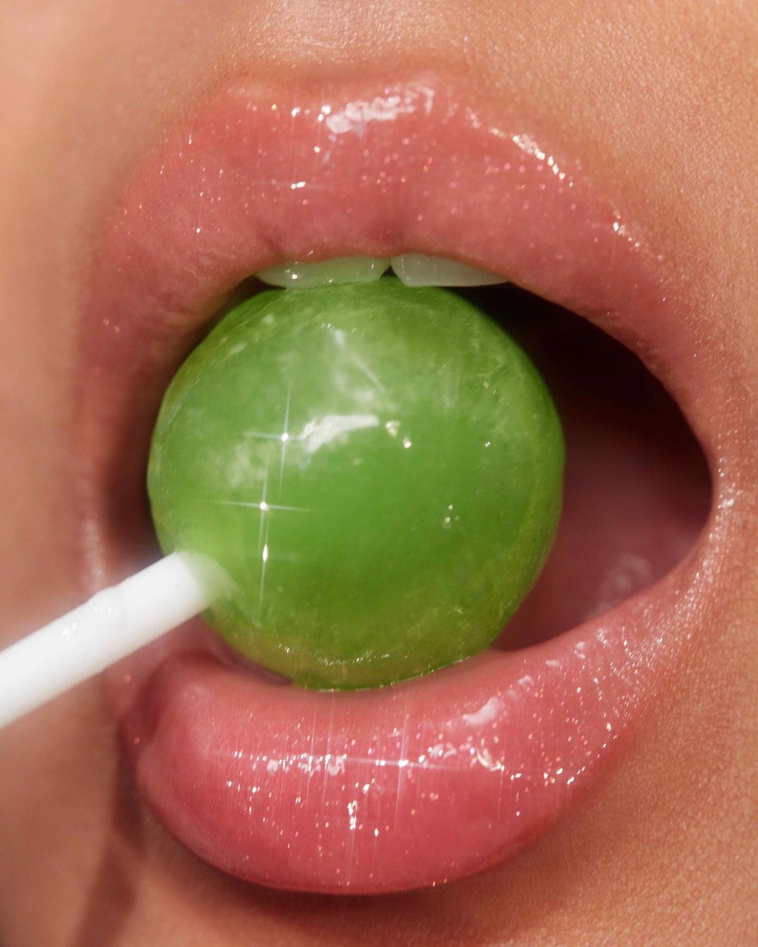 Close-up shot of a woman's glossy lips and teeth biting down on a green lollipop with a white stick, showcasing a vibrant beauty image.