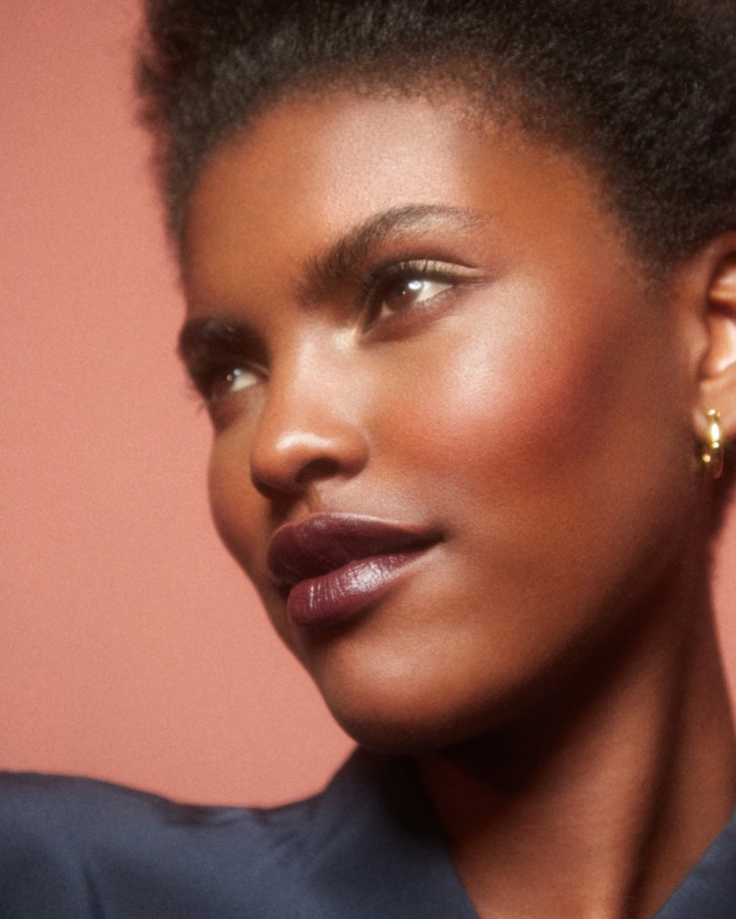 Close-up beauty portrait of a Black woman with dark lips and short dark hair wearing gold hoop earrings and a navy top.
