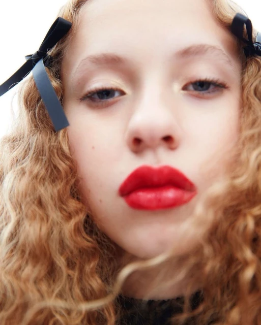 Close-up of a young person with curly blonde hair and red lipstick, wearing black ribbons in their hair, against a white background.