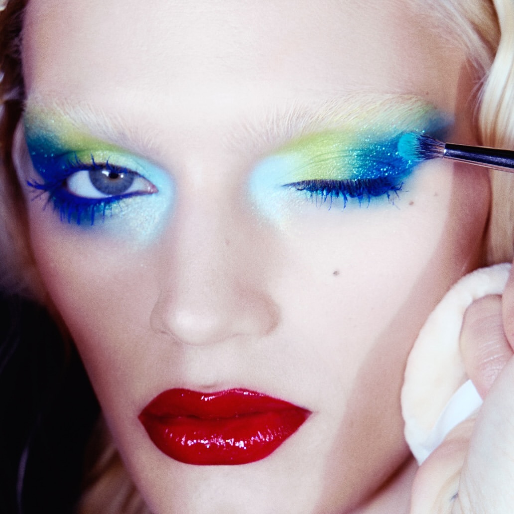 Close-up of a person's face with vibrant blue and green eye makeup being applied with a brush, emphasizing beauty and artistry.