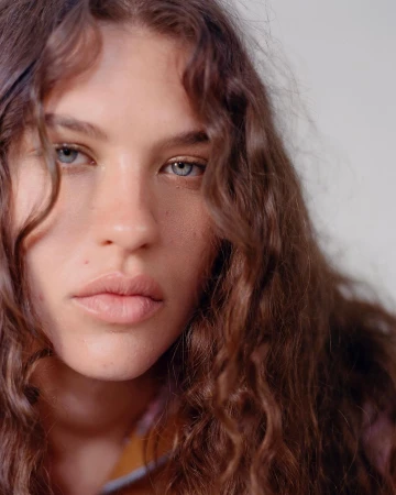 Close-up portrait of a woman with long wavy brown hair and blue eyes looking at the camera against a plain background