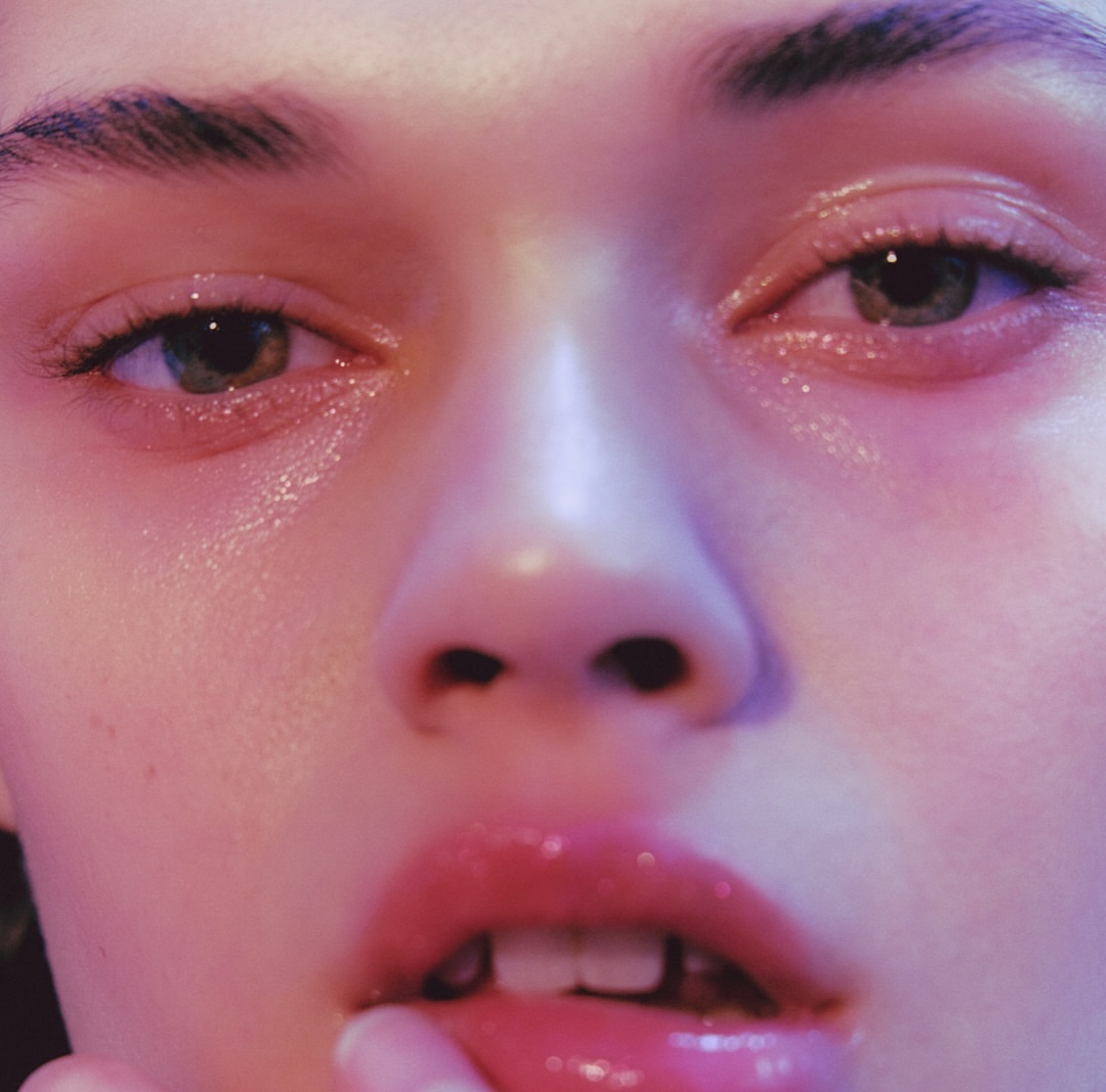 Close-up shot of an adult woman's face with glossy pink lips, green eyes, and pink lighting, with finger touching her lip.