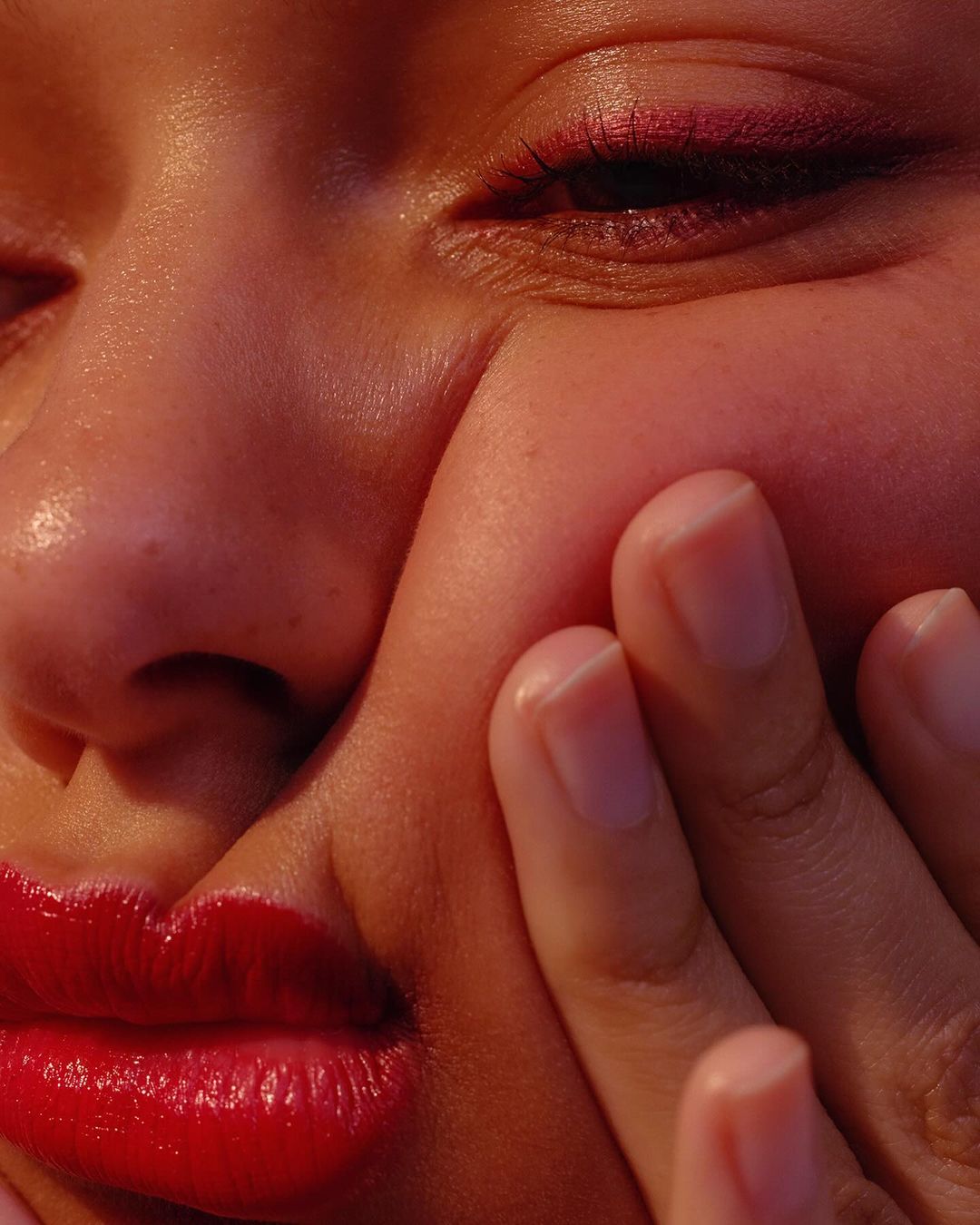 Close-up of a woman's face with red lipstick and her hand on her cheek. The focus is on her lips, eye, and hand with fingers.