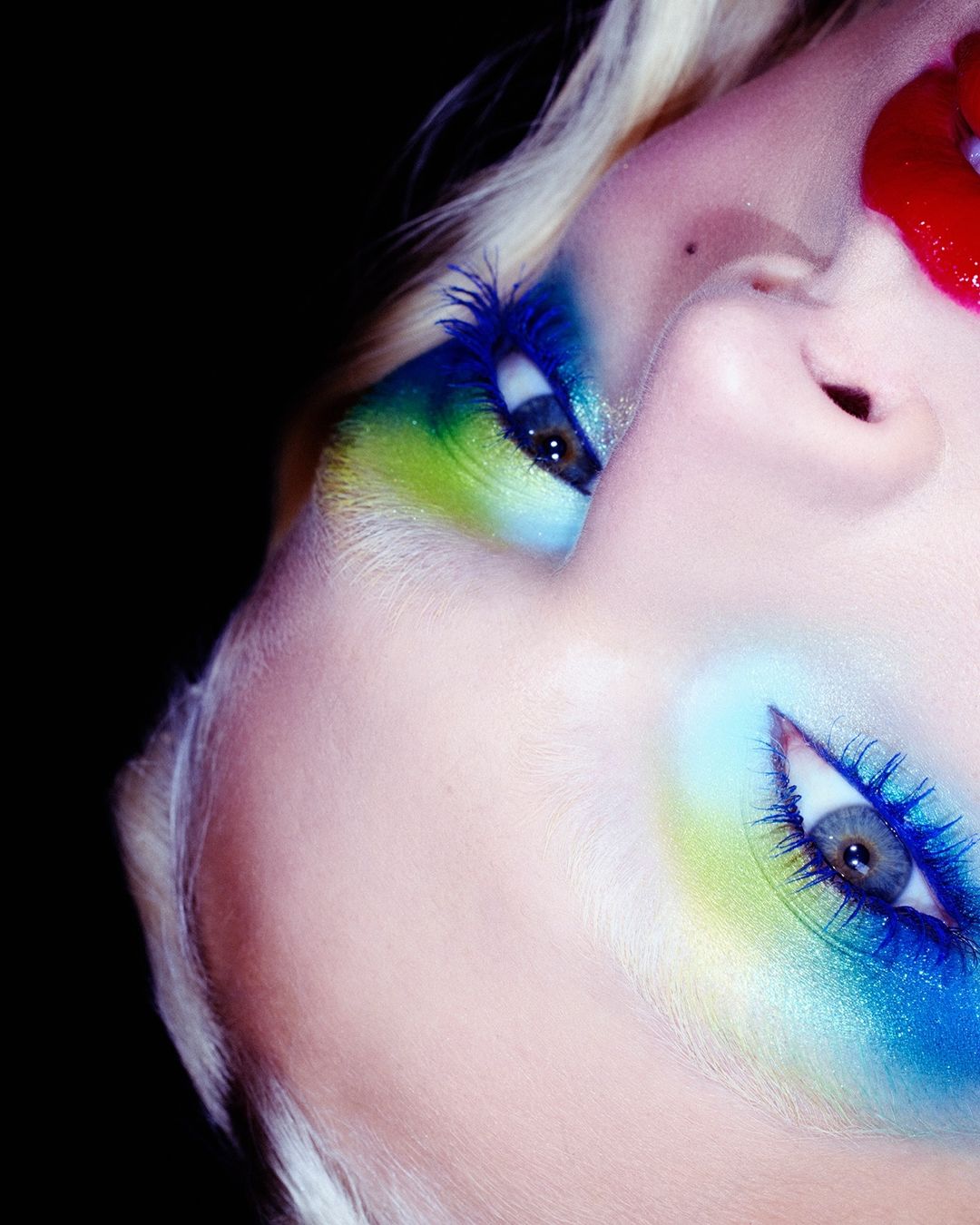 Close-up of a female face with vibrant blue and green makeup, showcasing a striking and artistic beauty look against a dark backdrop.