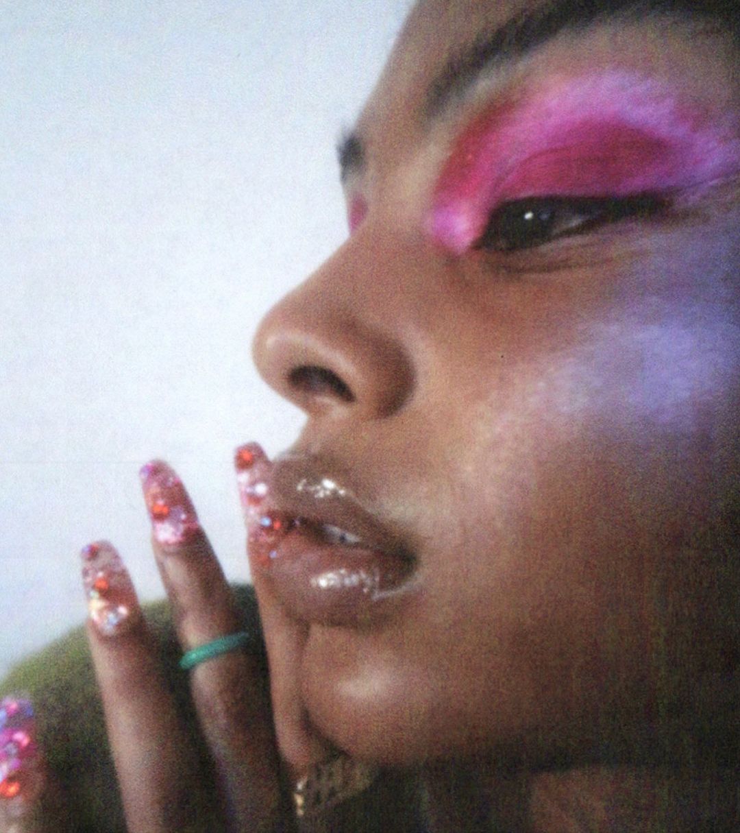 Close-up studio shot of a woman with pink eyeshadow and long, glittery nails, posing with her hand near her face against a bright background.