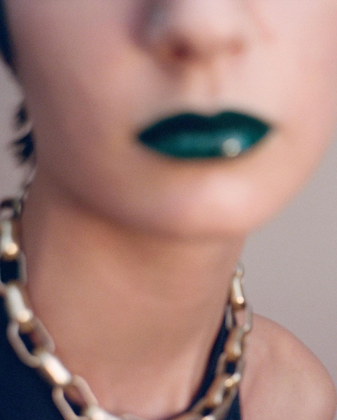 Close-up shot of an adult female with green lipstick and a thick gold chain around her neck, with a blurred background.