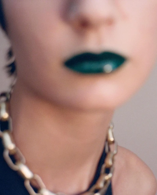 Close-up shot of an adult female with green lipstick and a thick gold chain around her neck, with a blurred background.