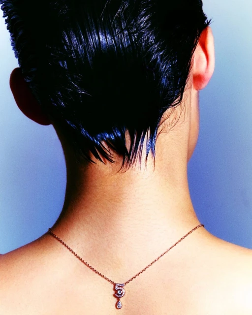 Back view of a woman with short, dark, wet hair, wearing a dainty gold necklace with a diamond pendant against a graduated blue background.