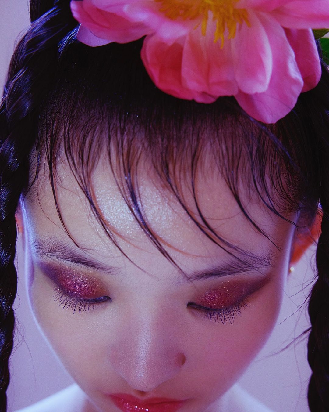 Close-up studio shot of a stylish female face with artistic eye makeup, pink lip gloss, and a large pink flower adorning her hair.