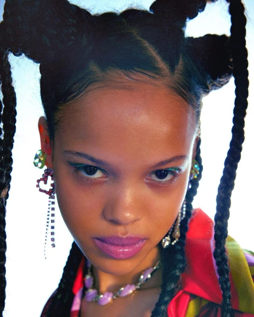 Close-up portrait of a young woman with braids, wearing colorful earrings and a lavender necklace, against a bright, neutral backdrop.
