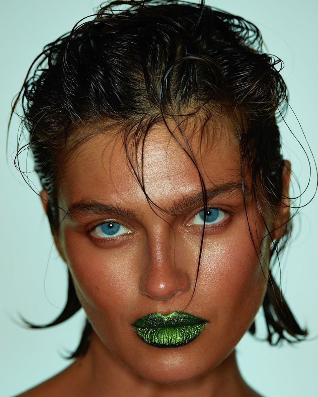 Close-up beauty shot of a young adult woman with wet dark hair, bright blue eyes, tan skin, and green glossy lipstick against a teal backdrop.