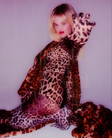 Woman in leopard print bodysuit and cape poses with outstretched hand against a light purple backdrop in a studio-style setup.