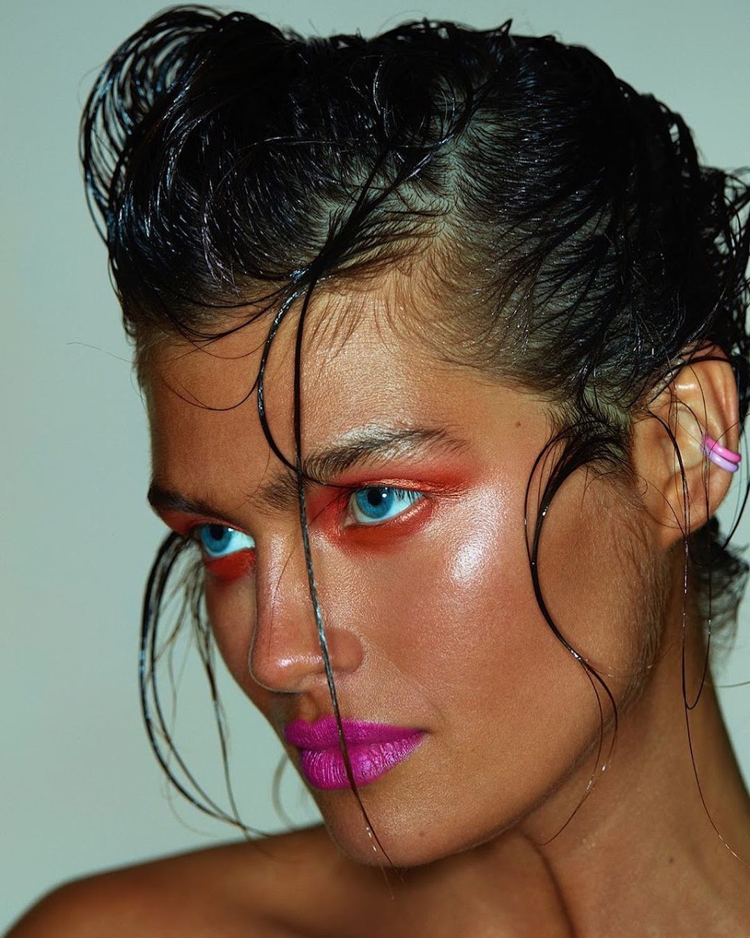 Close-up portrait of a woman with wet black hair, bright blue eyes, magenta lips, and red eye shadow against a neutral background.