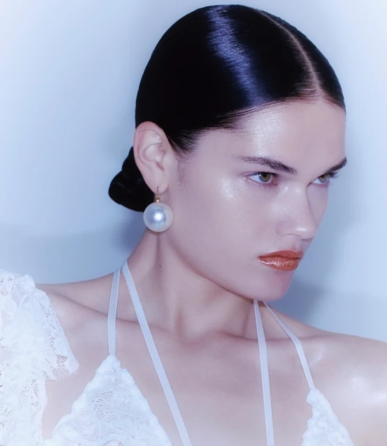 Close-up portrait of an adult female wearing a white lace top and pearl earring, against a soft blue background, with glossy hair and lips.